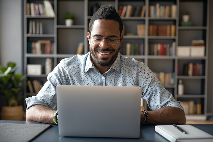 A smiling man using a laptop with a pen and pad by his side.