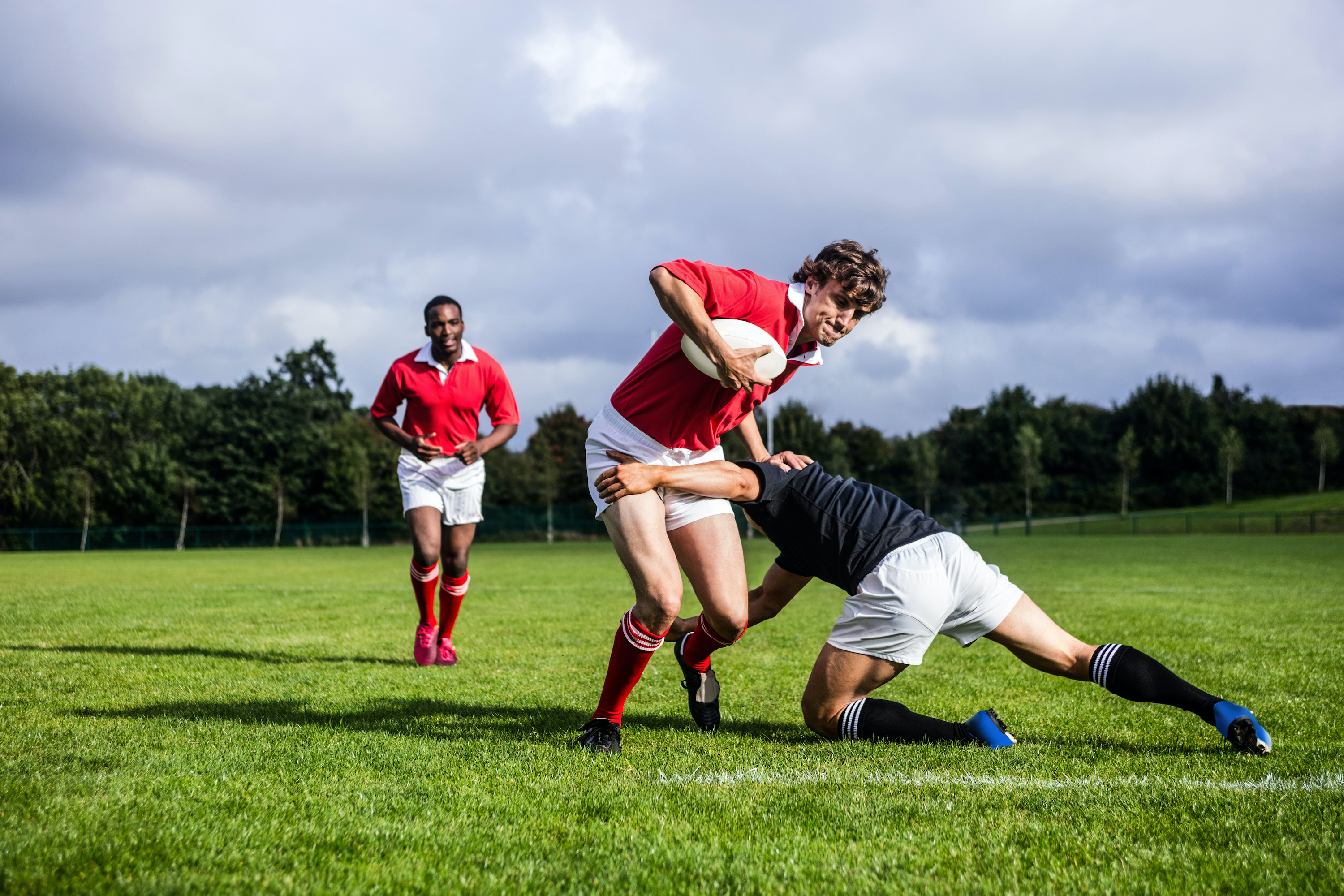 Rugby players on a field. The player at the front has the ball and is being tackled by a player from the opposing team.